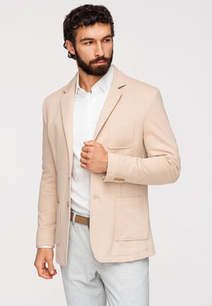 Homme aux cheveux bouclés foncés et à la barbe, portant un blazer beige, une chemise blanche et un pantalon gris clair, ajustant son blazer avec la main gauche.