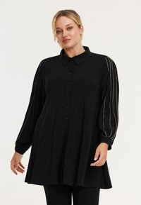 Zwarte button-up tuniek met een kraag en lange mouwen. Heeft zilveren accenten langs de mouwen, gemaakt van een zachte stof met een relaxte pasvorm.