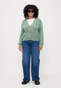 Light green knitted cardigan with a V-neck and wooden buttons, paired with wide-leg blue jeans and black clogs, featuring a textured design.