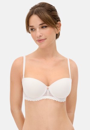 Mujer de piel clara y cabello castaño que lleva un sujetador blanco sin tirantes con detalles de encaje, mirando ligeramente hacia su izquierda sobre un fondo liso.