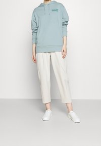 Sudadera de color azul claro con un bolsillo canguro y detalles de logo, combinada con pantalones recortados de color crema y zapatillas blancas. Tejido suave y casual.