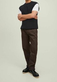 Schwarzes, geripptes, ärmelloses Stricktop über einem weißen T-Shirt, kombiniert mit dunkelbraunen Hosen und schwarzen Schnürschuhen mit einer beigen Sohle.