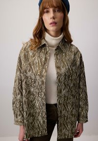 Camicia verde oliva a fantasia con disegni geometrici bianchi, con bottoni sul davanti e un fit rilassato, indossata sopra un maglione a collo alto crema.