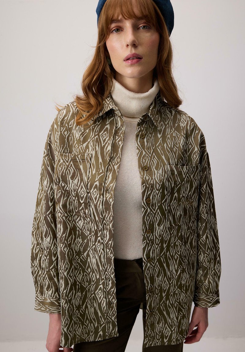 Camicia verde oliva a fantasia con disegni geometrici bianchi, con bottoni sul davanti e un fit rilassato, indossata sopra un maglione a collo alto crema.