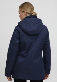 Veste imperméable bleu marine avec capuche, en tissu texturé, dotée de poches latérales et de poignets à boutons, vue de dos.