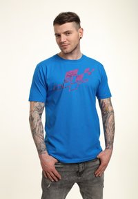 Modrá bavlněná tričko s fialovým grafickým designem na přední straně. Model nosí džíny s oprýskaným vzhledem a má tetování na pažích.