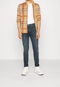 Randig knapp-up skjorta för män i tan och brunt, tillsammans med mörkblå jeans och vita sneakers. Skjortan har en krage och bröstfickor.