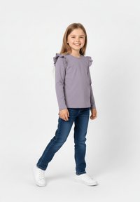 Długa koszula w jasnofioletowym kolorze z falbankami na ramionach, zestawiona z niebieskimi dżinsami i białymi tenisówkami; model stoi na neutralnym tle.