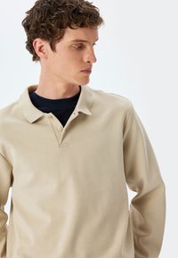 Beige pitkähihaine polo-paita, jossa on klassinen kaulus, kaksinappinen halkio ja teksturoitu puuvillakangas. Ominaisuuksia ovat hienovaraiset ompeluyksityiskohdat.