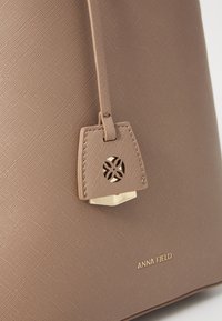 Brączowa torba o teksturowanej powierzchni, z złotym akcentem, małą metką z wyciętym wzorem i wytłoczonym napisem "ANNA FIELD" na powierzchni.