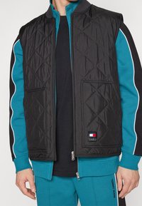 Svart vadderad väst med dragkedja fram, sidofickor och en Tommy Jeans-logotyp. Bärs över en teal-färgad träningsdräkt med svarta detaljer.