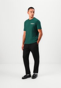 Grünes Baumwoll-T-Shirt mit Rundhalsausschnitt und kurzen Ärmeln, kombiniert mit schwarzen Hosen und schwarzen Schuhen, die ein dezentes Logo-Detail aufweisen.
