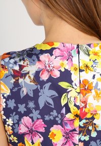 Blommig klänning med ett livfullt mönster av rosa, gula och blå blommor på en mörknavy bakgrund, samt en diskret bakre dragkedjeslutning.
