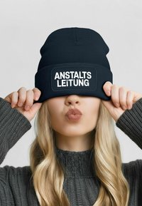 Black beanie with a white patch displaying the text "ANSTALTS LEITUNG." The wearer has long, wavy hair and is partially obscured by the hat.