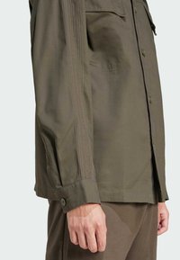 Camicetta verde oliva con una vestibilità strutturata, caratterizzata da due tasche frontali e dettagli in cucitura verticale lungo le maniche.