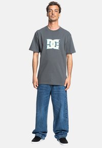 Grå bomullströja med en turkos logotyp på bröstet, matchad med löst sittande blå denimjeans och svarta slip-on skor.