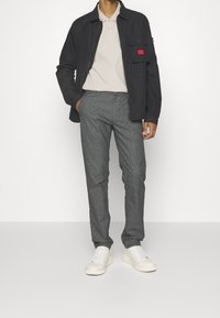 Black jacket with front pocket, beige polo shirt, gray patterned trousers, and white sneakers. Relaxed fit, smooth textures, and neutral colors.