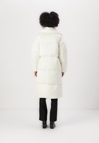 Hvit puffjakke med lang design, høy krage og quiltet tekstur. Kombinert med svarte bukser og kraftige hæleboots.