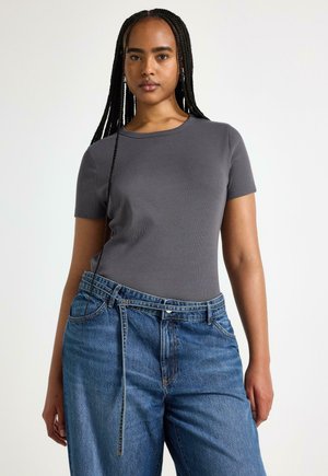 Ung kvinde med langt fletning og iført en tætsiddende mørkegrå t-shirt med korte ærmer og højtaljede blå jeans med bælte.
