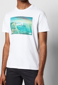 En person som bär en vit T-shirt med en blå och grön landskapsgrafik som har texten "Levi's" står framför en enfärgad bakgrund.