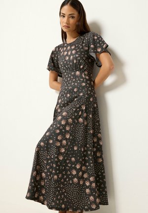Kvinde med langt brunt hår iført en sort blomster maxi-kjole med flagrende ærmer, stående mod en hvid væg.