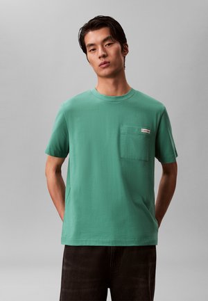 Junger Mann mit dunklem Haar trägt ein grünes kurzärmeliges T-Shirt mit Brusttasche und kleinem Calvin Klein-Label, steht vor einem grauen Hintergrund.