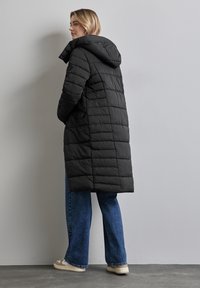 Long manteau doudoune noir avec un motif matelassé horizontal et une capuche, associé à un jean bleu à jambes larges et des sneakers claires.