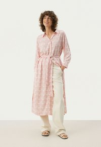 Chemise longue rose clair avec un motif floral, devant boutonné, taille ceinturée, manches longues, assortie à un pantalon beige clair et des sandales.