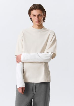 Młody mężczyzna o średniej długości włosach, ubrany w warstwowo kremowo-białe koszule z długim rękawem oraz szare spodnie, stojący z jedną ręką skrzyżowaną na ciele.