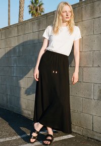 Jauna sieviete ar gariem blondiem matiem atrodas pie betona sienas svaigā gaisā, valkājot baltu T-kreklu, melnu maksi svārku un melnas sandales.