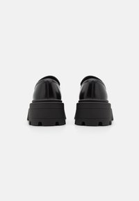 Zwarte patenten leren schoenen met een ronde vorm en dikke zwarte rubberen zolen, die een gestructureerd oppervlak en een uitgesproken profielpatroon hebben.