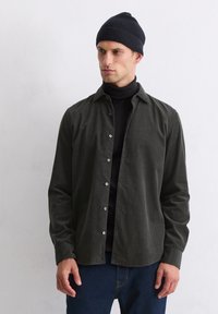 Chemise boutonnée tissée vert foncé avec un col, des manches longues et des boutons semblables à des perles, portée sur un col roulé noir, associée à un jean bleu.