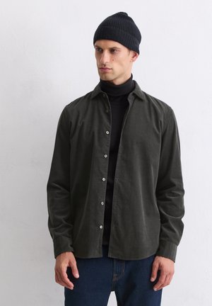 Dark green woven button-up shirt with a collar, long sleeves, and pearl-like buttons, worn over a black turtleneck, paired with blue jeans.