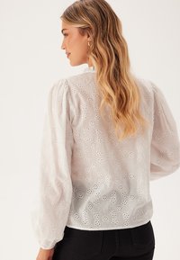 Witte, lichte blouse met opgezette mouwen, voorzien van gehaakte bloemenpatronen. De achterkant heeft een ronde halslijn en verfijnde stikdetails.