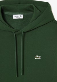 Sudadera Lacoste de color verde oscuro con cordones y pequeño logo de cocodrilo bordado en el pecho, etiquetada como ajuste clásico.