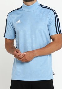 Ljusblå Adidas t-shirt med kort ärm och svarta axelränder samt subtilt mönstrad textur, bärs av person med hopknäppta händer.