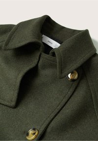Kabát z vlněné směsi olivově zelené barvy s širokým límcem, dvojřadým designem a knoflíky ve zlatém tónu. Má měkkou texturu a strukturovaný tvar.