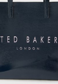 Tumesinise tekstuuriga õlakott, millel on "TED BAKER LONDON" kaubamärgi logo reljeefsete hõbedaste tähtedega ning kaks lühikest käepidet.