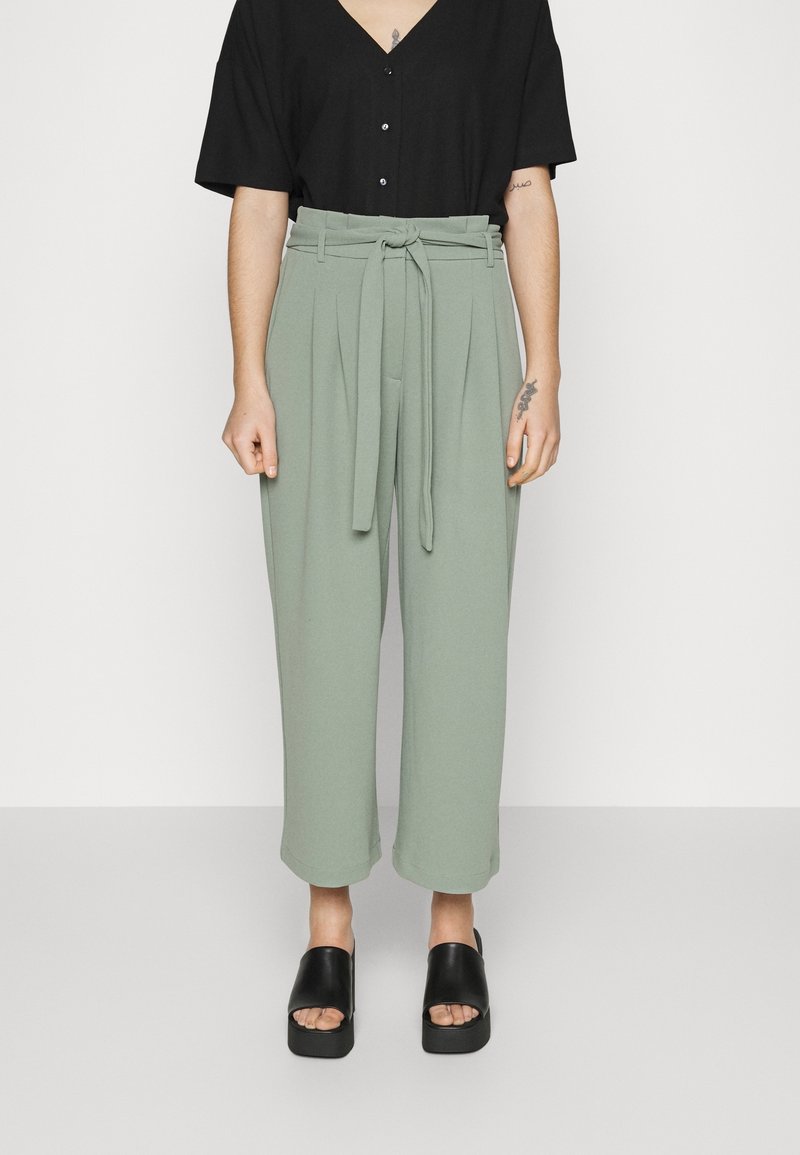 Pantalon vert taille haute avec ceinture, design plissé et jambes larges, associé à une chemise noire à col en V boutonnée et des sandales compensées noires.