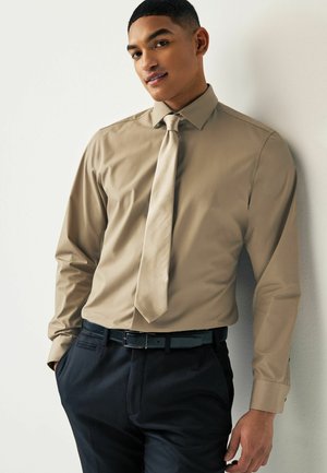 Camicia beige a maniche lunghe con cravatta abbinata, abbinata a pantaloni scuri e una cintura nera; tessuto liscio, design aderente, lucentezza sottile.
