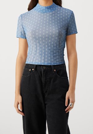 Kvinde iført en tætsiddende blå kortærmet top med et gentagende "HUGO" logo mønster kombineret med sorte høj taljede jeans.
