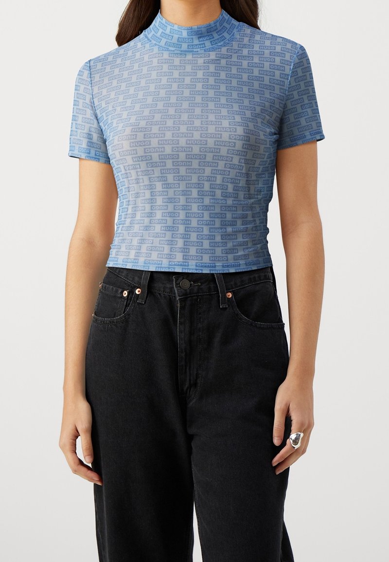 Femme portant un top bleu ajusté à manches courtes avec un motif répétitif du logo "HUGO", associé à un jean noir taille haute.