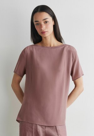 Kobieta z długimi ciemnymi włosami, ubrana w mauve krótką bluzkę i pasujące spodnie, stojąca przy jasnoszarej ścianie.