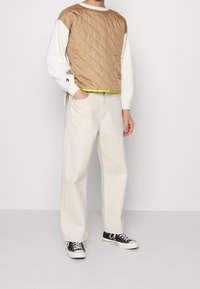 Chaleco acolchado marrón sobre una camiseta de manga larga blanca, combinado con pantalones blancos holgados y zapatillas negras. Tiene un ribete verde neón.