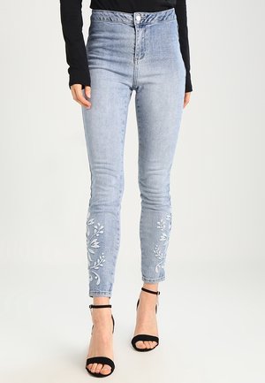 Hellblaue Skinny-Jeans mit hoher Taille und weißer Blumenstickerei auf den unteren Beinen, kombiniert mit einem schwarzen Langarmtop und schwarzen offenen High Heels.