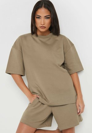 Kobieta z prostymi ciemnymi włosami, ubrana w oversize beżowy t-shirt i dopasowane szorty, stojąca z jedną ręką w kieszeni na jednolitym tle.