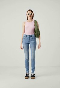 Ženska v roza brezrokavniku, svetlih modrih skinny kavbojkah, črnih športnih copatih, belih sončnih očalih, s zeleno torbico na rami, stoji pred enobarvno ozadjem.