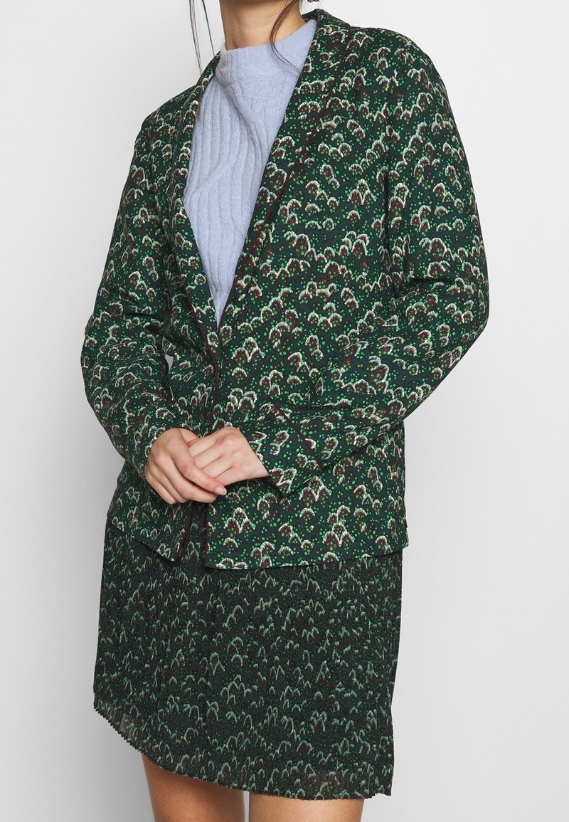 Femme portant un blazer vert à motif fleuri et une jupe assortie, avec un pull côtelé bleu clair en dessous, les mains jointes devant.