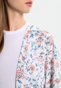 Květinové kimono z lehké bílé tkaniny s růžovými a modrými květinovými vzory, otevřeným předním designem a krátkými rukávy.