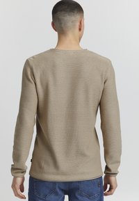 Beige Langarmhemd mit einem strukturierten, gewebten Muster und Rundhalsausschnitt. Integriertes Etikett an der Taille, kombiniert mit blauen Jeans.